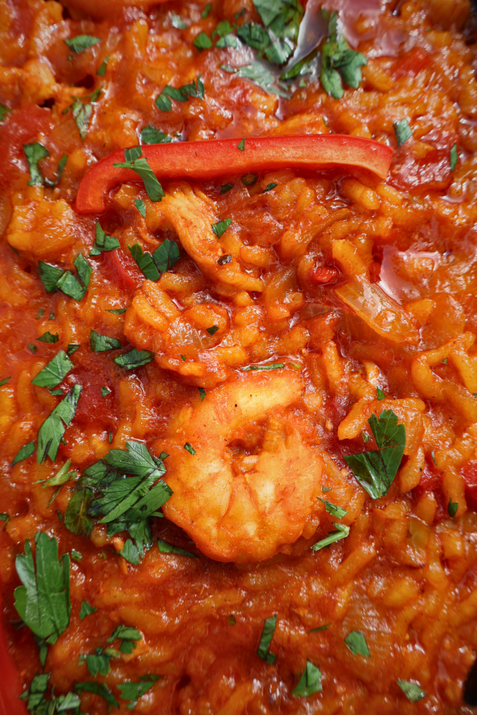 Close up of tomato paella rice with shrimp, red bell pepper strips, and fresh parsley, showing the rich orange color from sazon con achiote