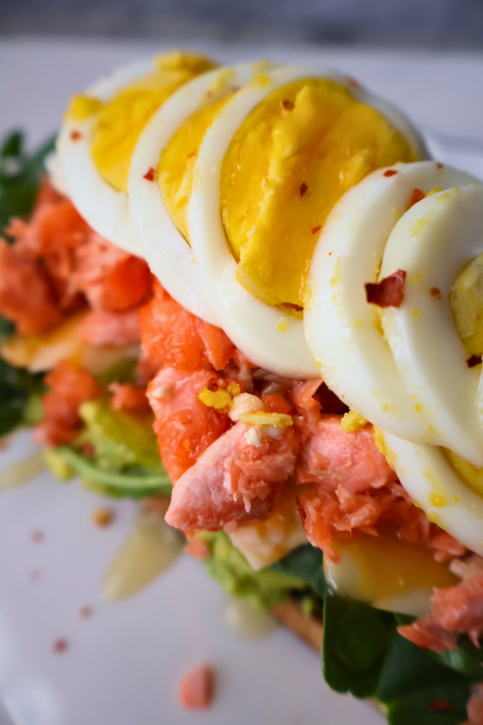 Angled side view of open faced salmon and avocado sourdough sandwich on a white scalloped gold rimmed plate showing layers of mashed avocado, arugula, flaked fresh salmon, and fanned boiled egg slices shot in natural light.