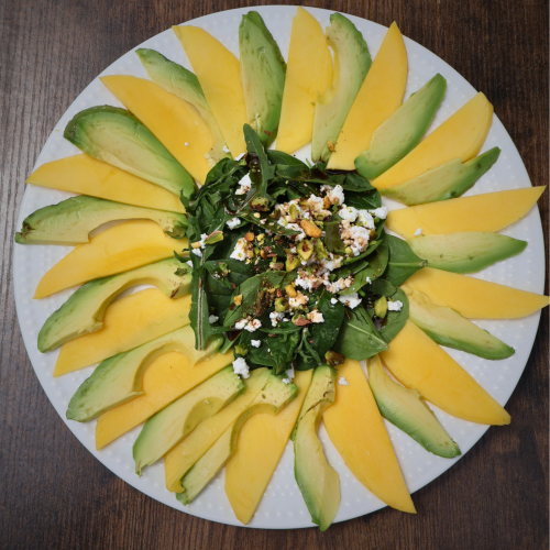 Mango and avocado carpaccio salad arranged in a sunflower pattern on a white round plate, topped with arugula, spinach, Bulgarian feta crumbles, chopped pistachios, and pomegranate molasses