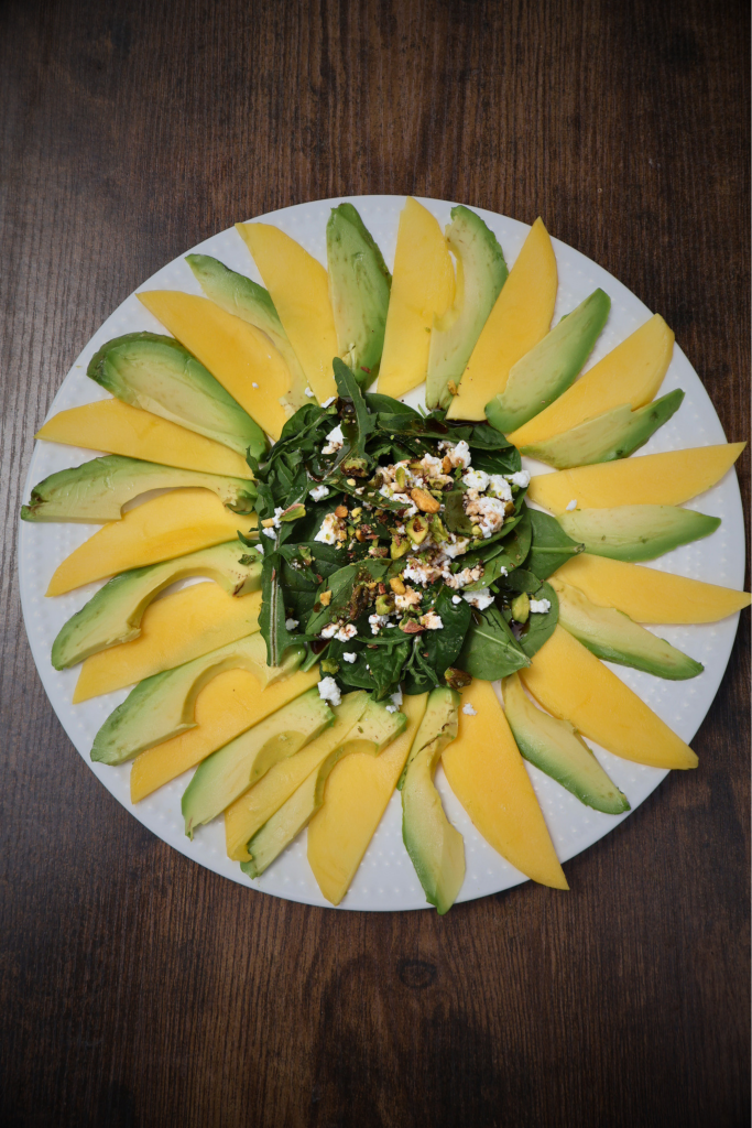 Mango and avocado carpaccio salad arranged in a sunflower pattern on a white round plate, topped with arugula, spinach, Bulgarian feta crumbles, chopped pistachios, and pomegranate molasses