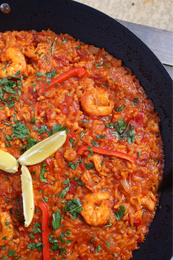 Wide angle shot of a full black paella pan with tomato rice, chicken chunks, shrimp, and red bell pepper, garnished with fresh parsley and lemon wedges, photographed outdoors on a wooden surface.