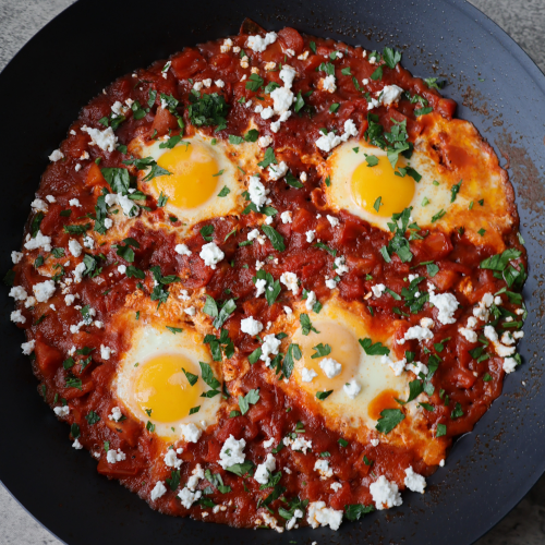 Shakshuka with four poached eggs in a rich red tomato sauce topped with Bulgarian feta crumbles and fresh chopped parsley served in a large black pan on a gray surface