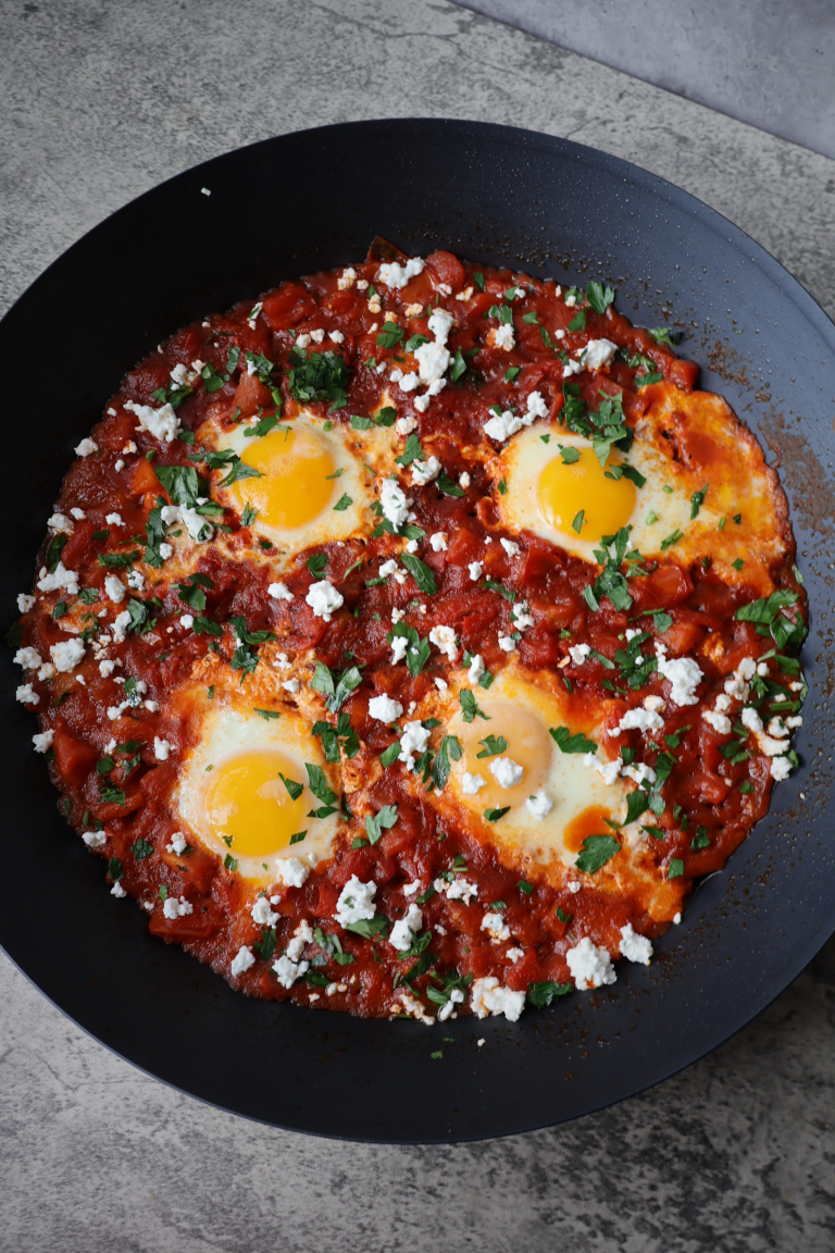Shakshuka with four poached eggs in a rich red tomato sauce topped with Bulgarian feta crumbles and fresh chopped parsley served in a large black pan on a gray surface