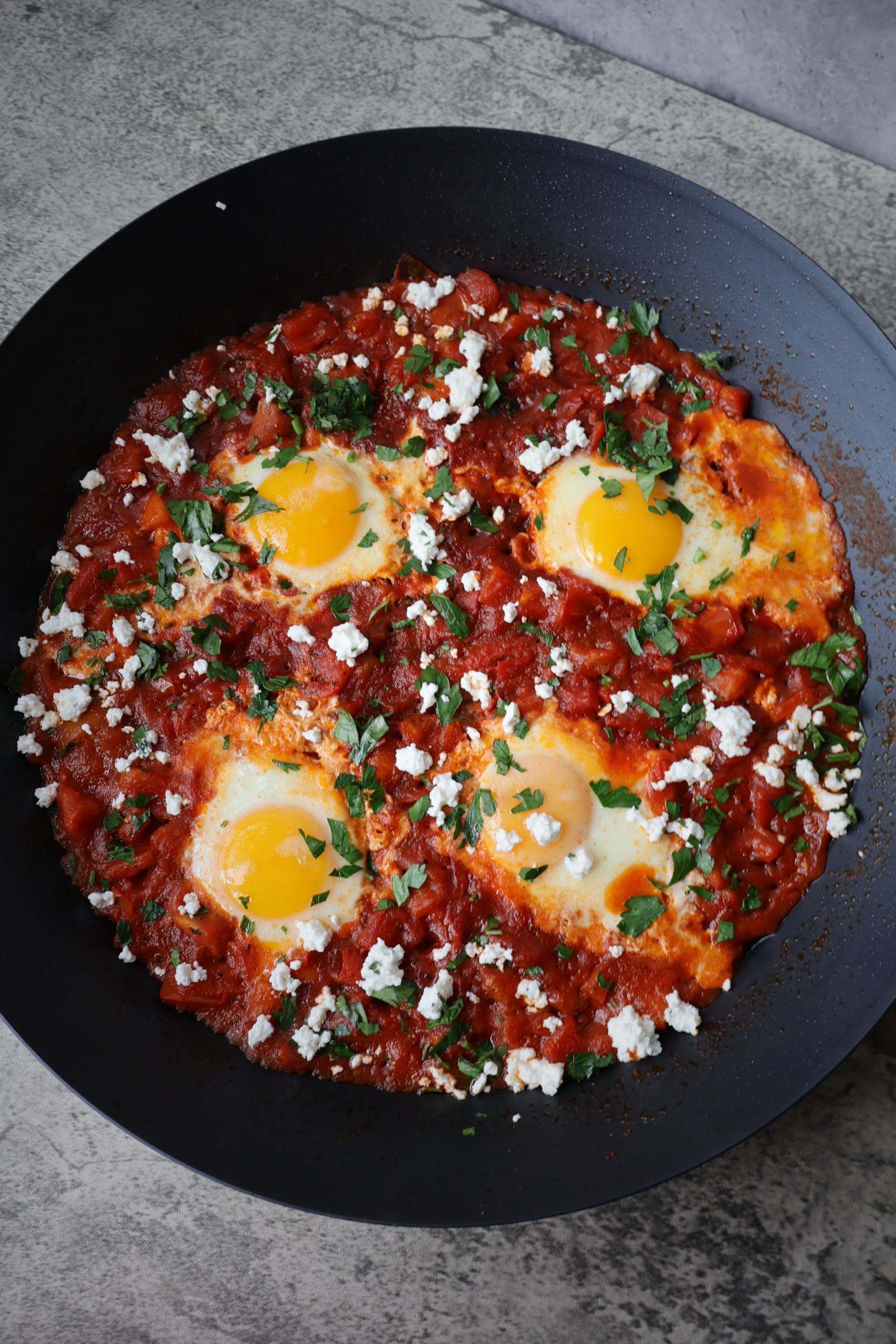 Shakshuka with four poached eggs in a rich red tomato sauce topped with Bulgarian feta crumbles and fresh chopped parsley served in a large black pan on a gray surface