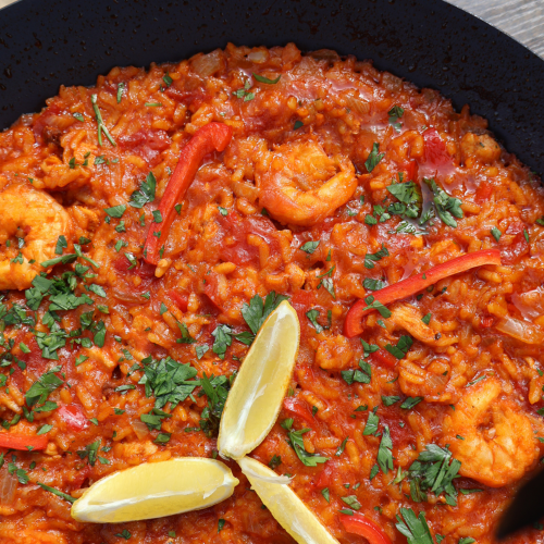 Tomato paella with chicken and shrimp in a large black paella pan, garnished with fresh parsley, lemon wedges, and red bell pepper strips, photographed from above