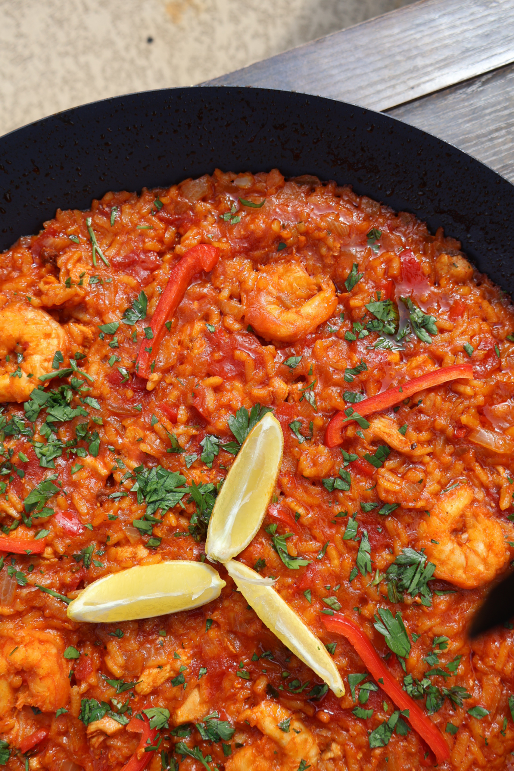 Tomato paella with chicken and shrimp in a large black paella pan, garnished with fresh parsley, lemon wedges, and red bell pepper strips, photographed from above