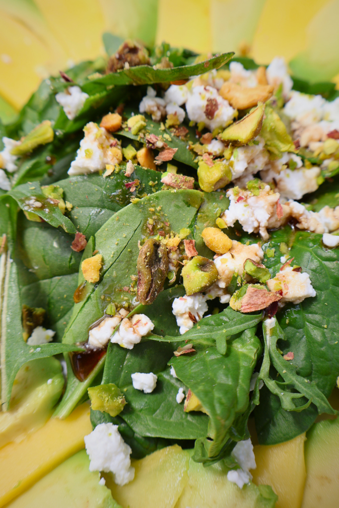 Close up of Mango and avocado carpaccio salad arranged in a sunflower pattern on a white round plate, topped with arugula, spinach, Bulgarian feta crumbles, chopped pistachios, and pomegranate molasses