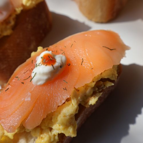 Close up of smoked salmon and Boursin scrambled egg baguette bite topped with a sour cream dot showing the layers of creamy eggs and silky smoked salmon on toasted baguette