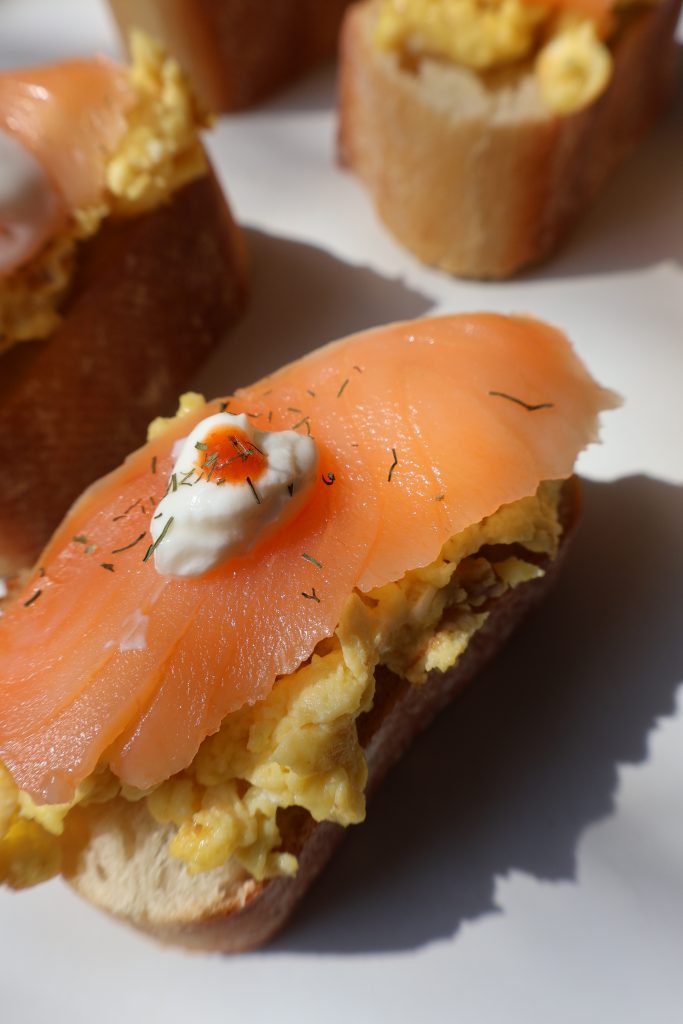 Close up of smoked salmon and Boursin scrambled egg baguette bite topped with a sour cream dot showing the layers of creamy eggs and silky smoked salmon on toasted baguette