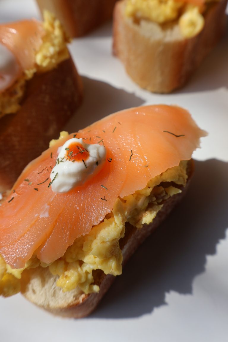 Close up of smoked salmon and Boursin scrambled egg baguette bite topped with a sour cream dot showing the layers of creamy eggs and silky smoked salmon on toasted baguette