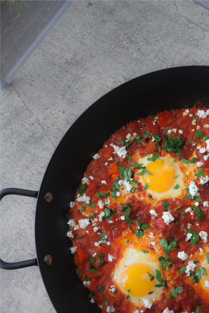 Shakshuka with poached eggs in tomato sauce topped with Bulgarian feta and fresh parsley in a black pan, photographed from a slight angle showing the full pan