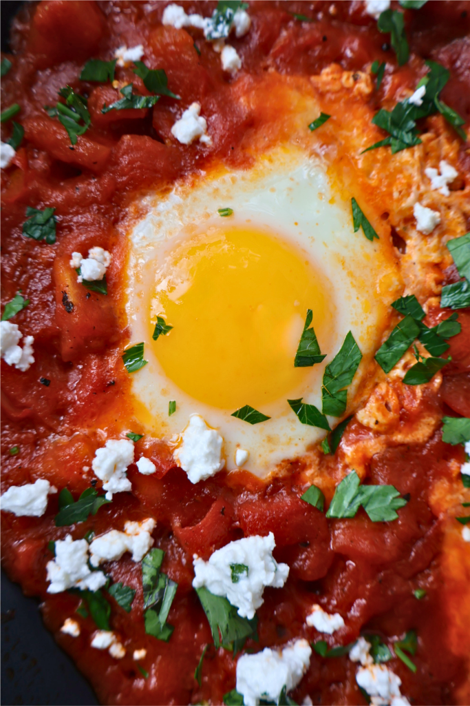 Close up of a single egg poached in rich red tomato sauce with Bulgarian feta crumbles and fresh parsley, from a shakshuka made with pomegranate molasses