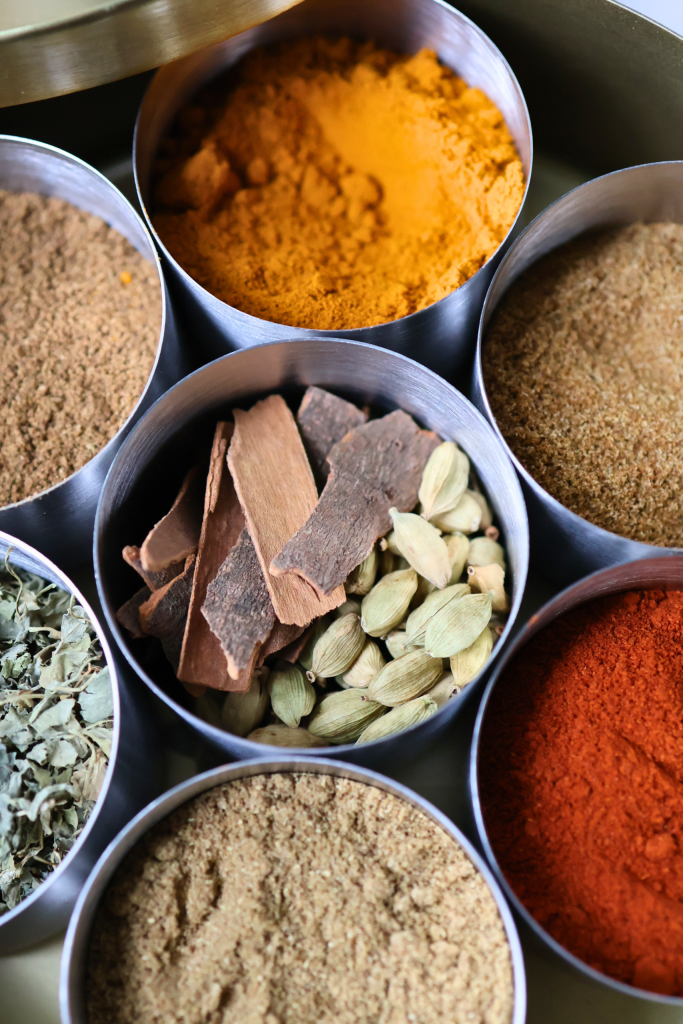 Overhead view of a masala dabba with seven spices including turmeric, red chili powder, cinnamon sticks, cardamom pods, and ground cumin and coriander