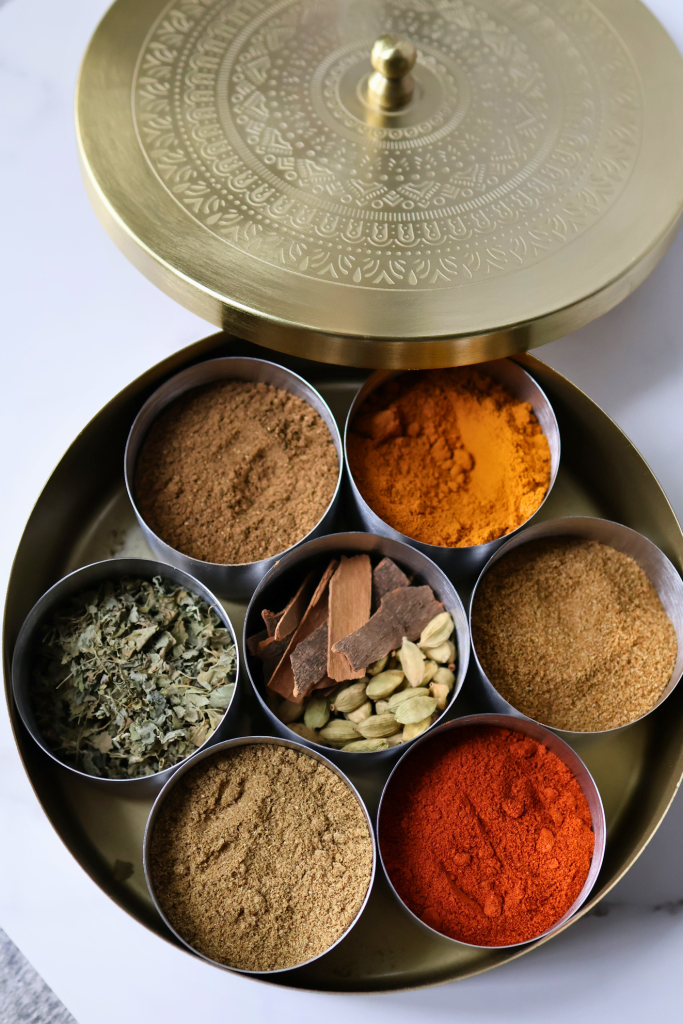 Gold masala dabba filled with Indian spices including turmeric, red chili powder, cinnamon, cardamom, and ground cumin