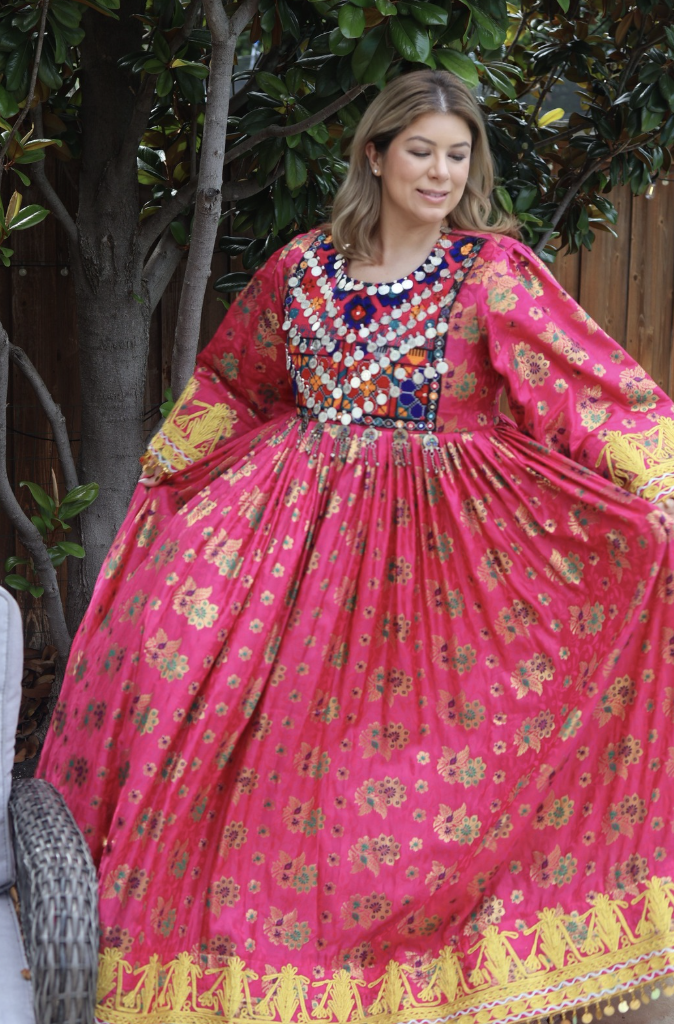 Woman wearing a traditional Afghan dress with intricate embroidery and mirrorwork details, standing outdoors and holding the flowing skirt during a cultural gathering.