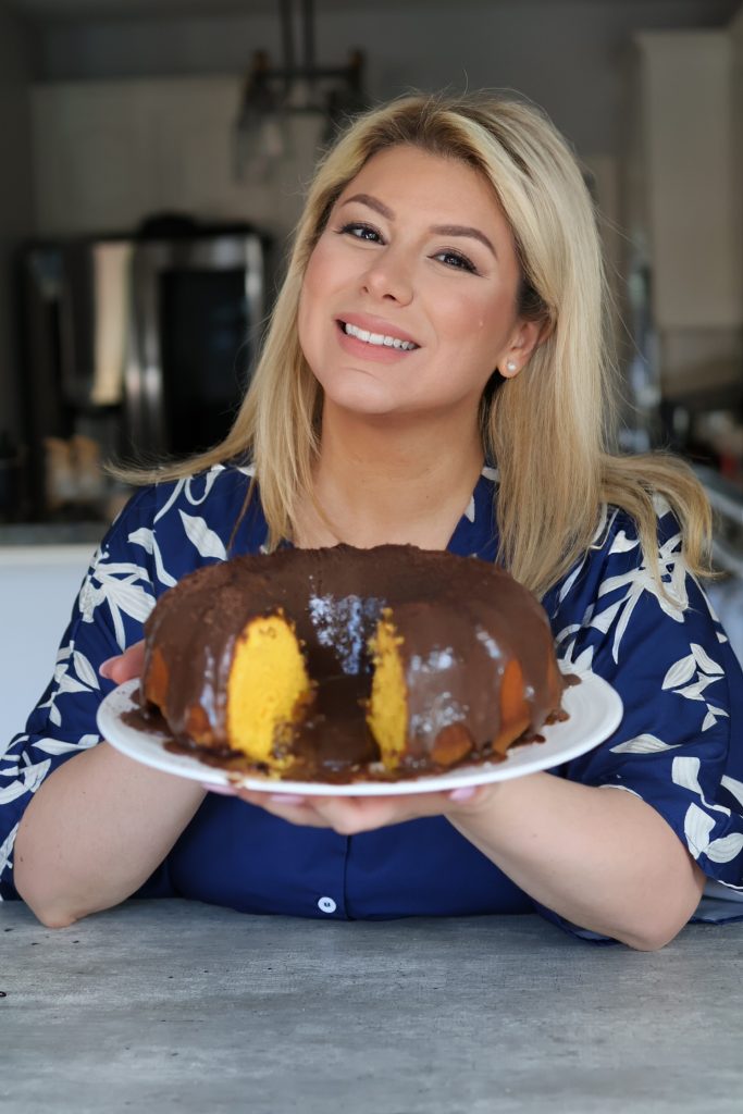 Smiling woman in a blue patterned dress holding a Brazilian carrot cake (bolo de cenoura) with chocolate glaze on a white plate