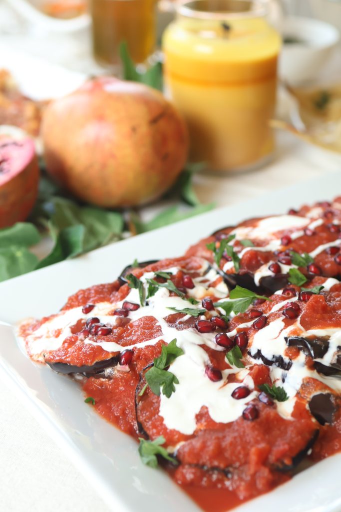 Traditional Afghan borani banjan made with tender eggplant, tomato sauce, garlic yogurt, and topped with fresh herbs and pomegranate seeds.