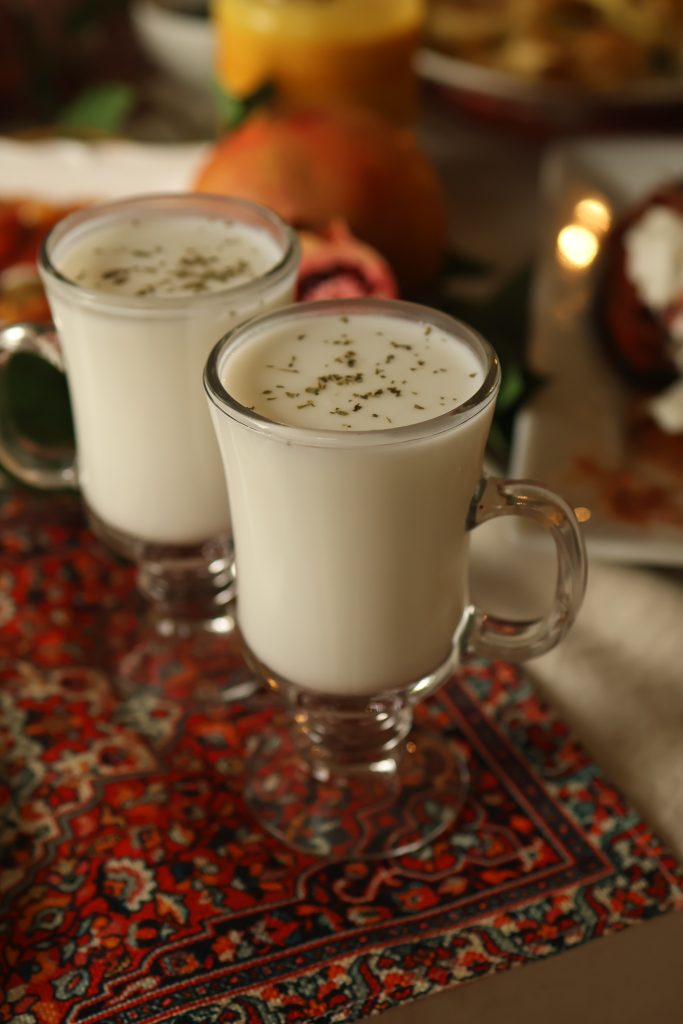 Two traditional glasses of Afghan doogh, a yogurt-based drink with dried mint, served alongside a spread of Afghan dishes.