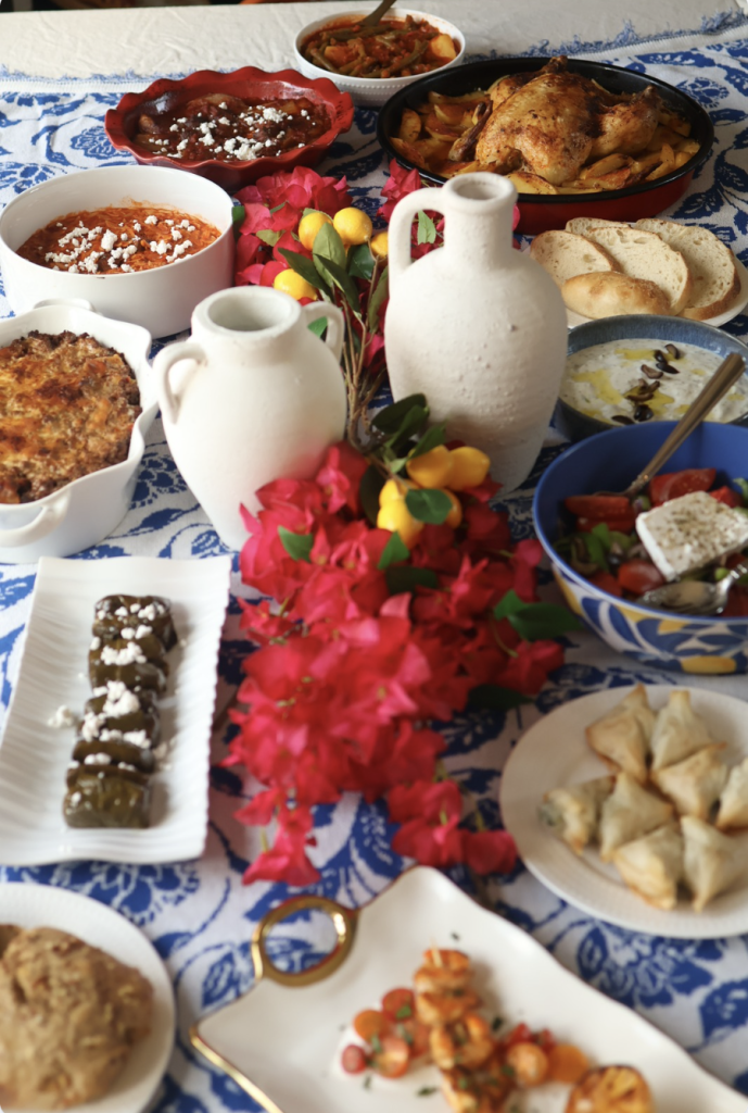 Greek table set family style with platters, dips, and bread around a centerpiece of flowers and lemons