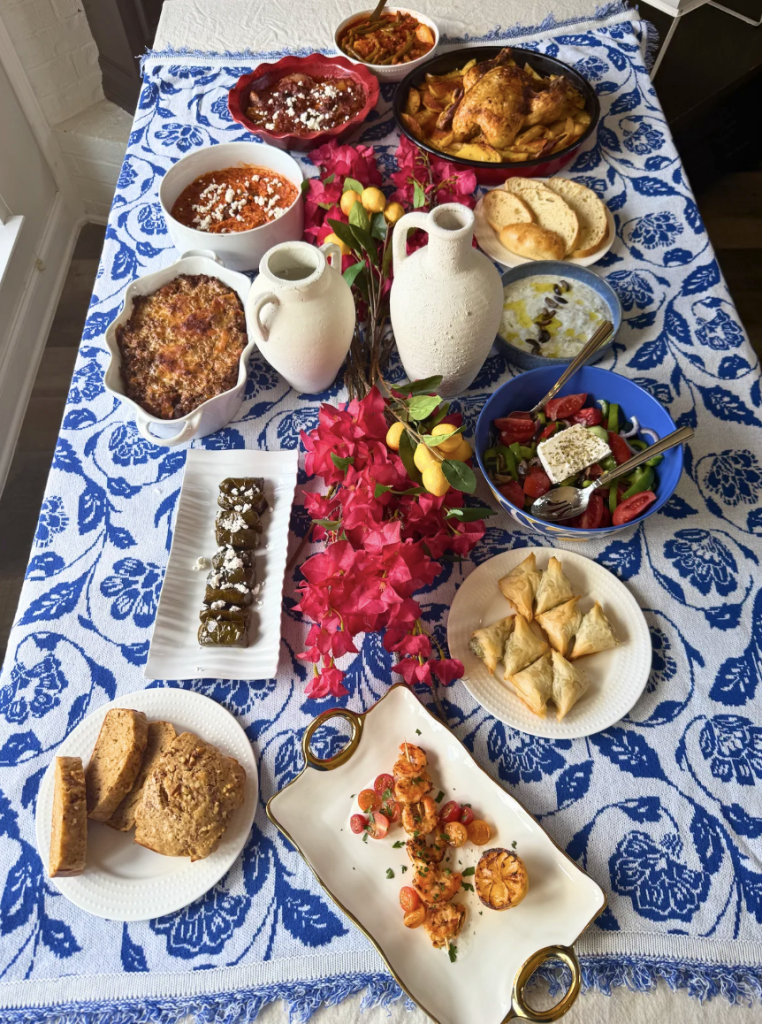 A full Greek table set on blue and white linen with shrimp souvlaki, Greek salad, bread, dips, and platters served family style