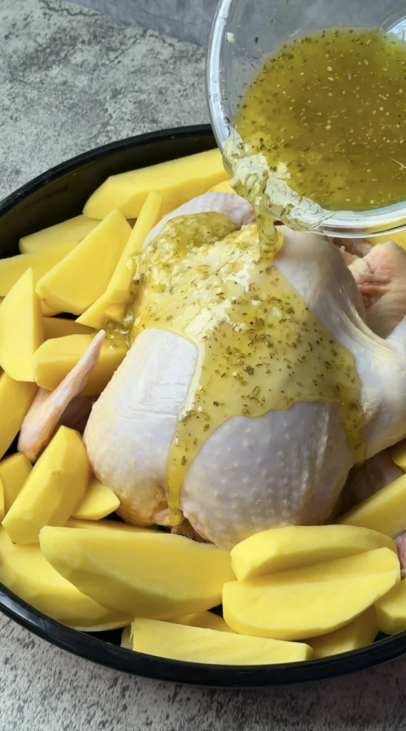 Raw chicken and potato wedges in a roasting pan with lemon oregano marinade being poured over for Greek chicken and potatoes