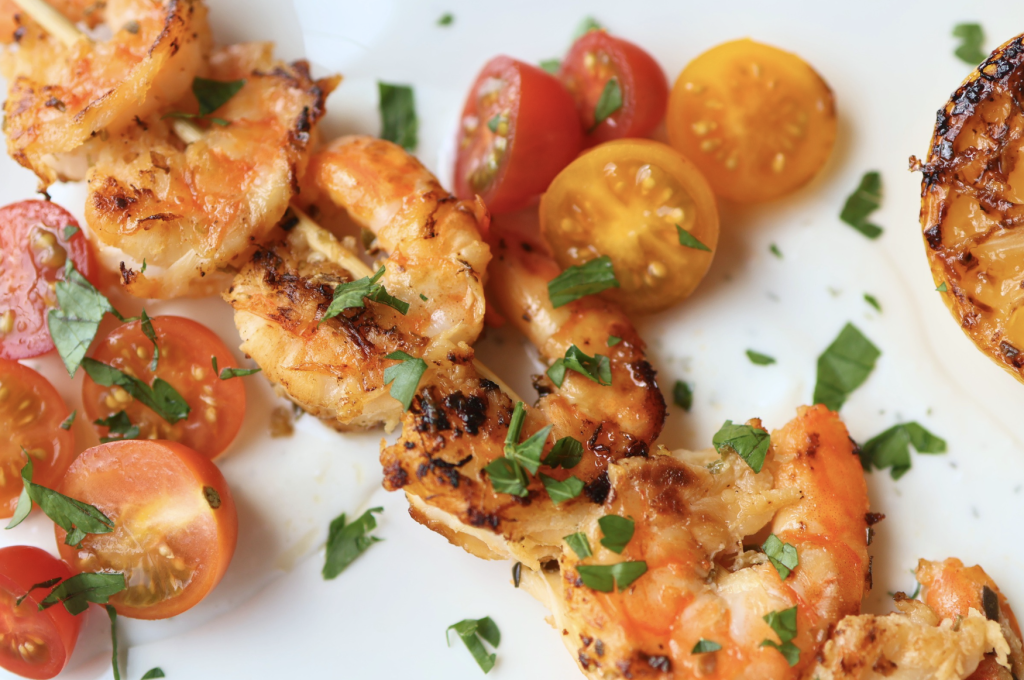 Grilled shrimp souvlaki with charred edges, cherry tomatoes, and fresh herbs on a white plate