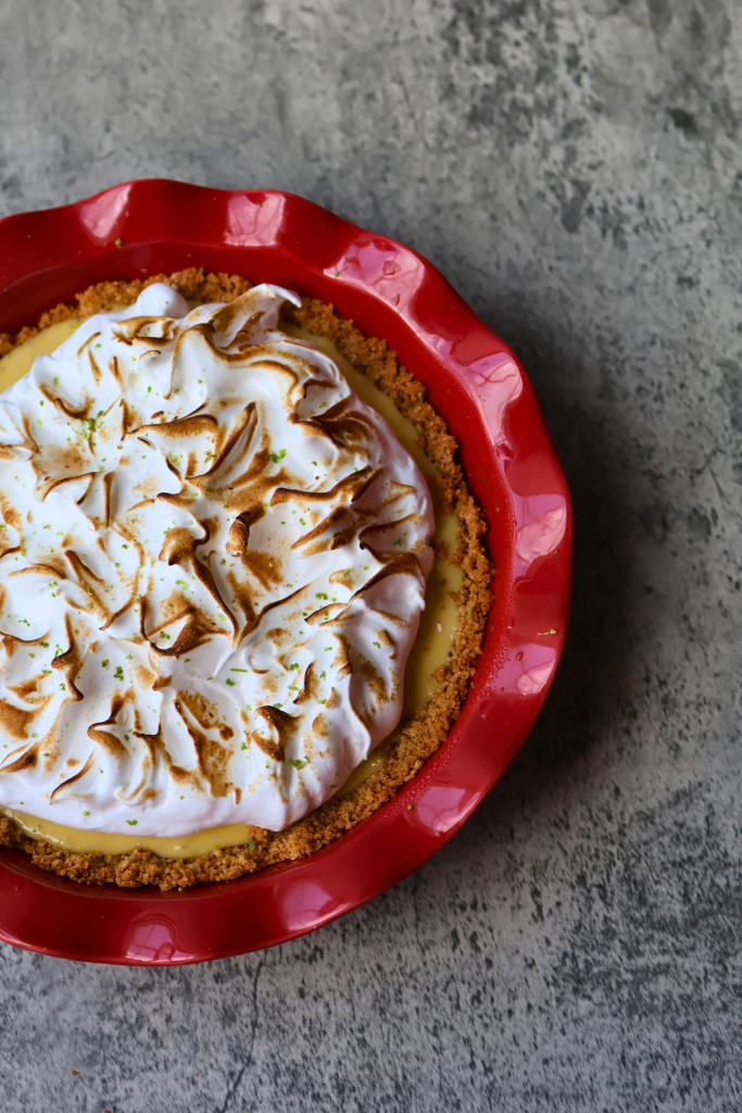 Overhead view of key lime pie with toasted meringue swirls and lime zest in a red ceramic pie dish on a gray surface.
