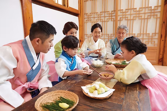 Korean family wearing hanbok gathered around a table sharing food during a traditional celebration.