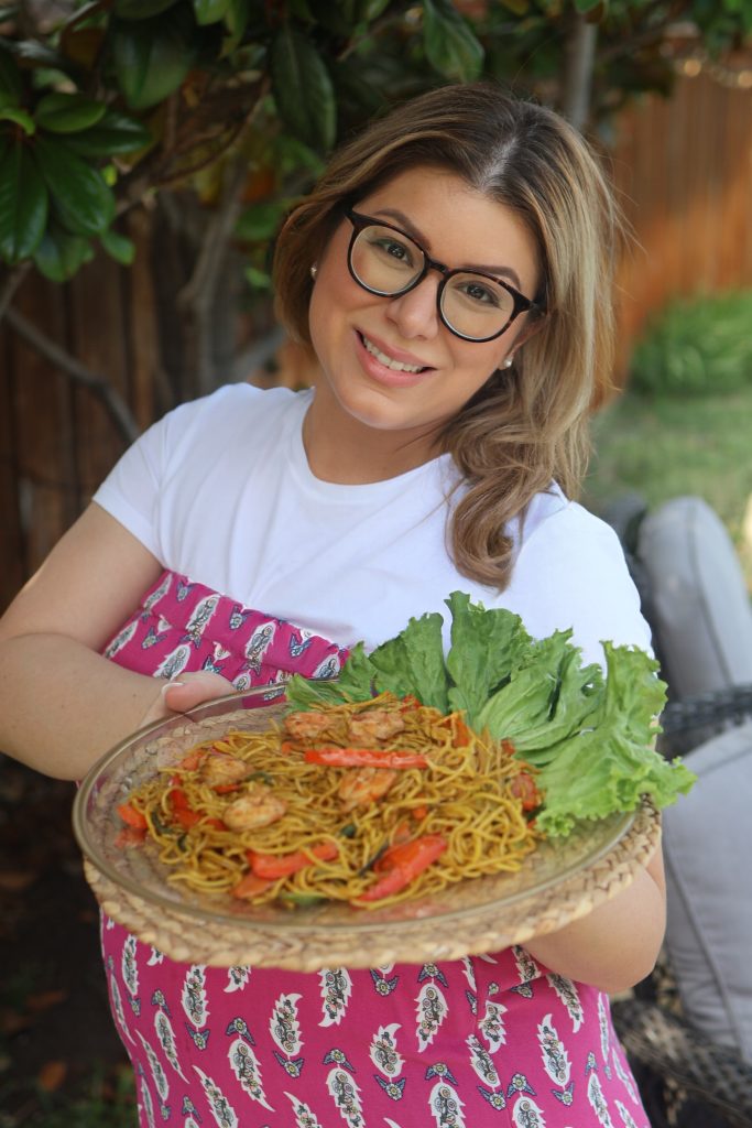 Emilia Kim holding a platter of traditional Malagasy mine sao, a stir-fried noodle dish with shrimp and vegetables, while hosting Madagascar for her Hosting the World series.