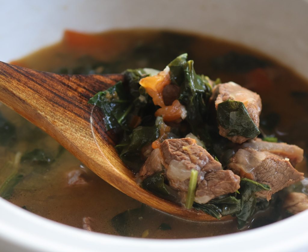 Romazava, Madagascar’s national dish, served in a white bowl with a wooden spoon, featuring tender beef and leafy greens in a rich savory broth.