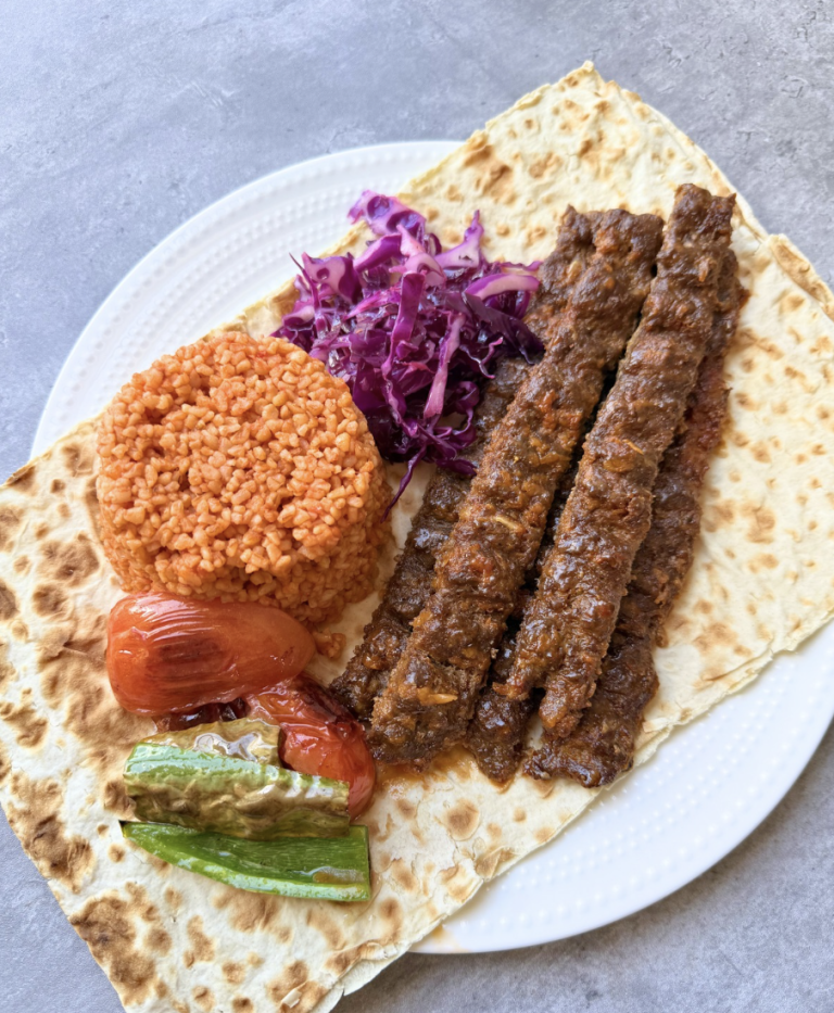 Turkish Adana kebab served with bulgur pilaf, lavash bread, red cabbage salad, grilled tomato, and green pepper.