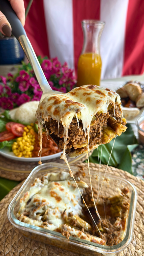Cheesy Puerto Rican pastelón made with sweet plantains, seasoned ground beef, and melted cheese being lifted from a baking dish with long, stretchy cheese pulls.