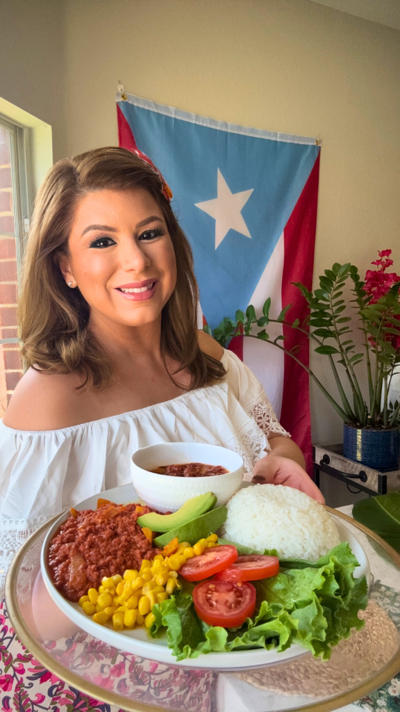 Emilia Kim holding a traditional Puerto Rican plate with white rice, stewed beans, Puerto Rican corned beef, avocado, sweet corn, lettuce, and tomatoes in front of a Puerto Rican flag.