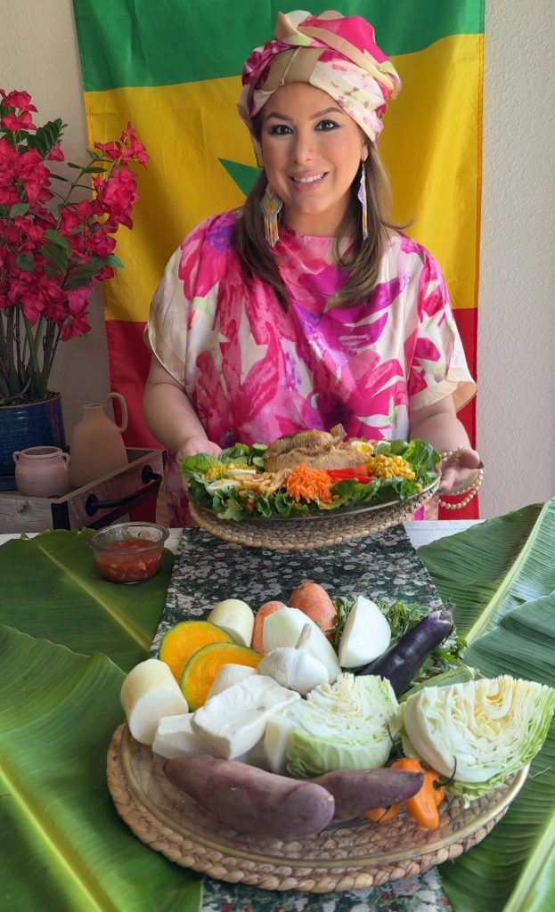 Woman wearing traditional Senegalese-inspired clothing seated behind a colorful Senegalese table featuring thieboudienne, fresh vegetables, eggs, cabbage, and banana leaf decorations as part of a Hosting the World cultural dinner.
