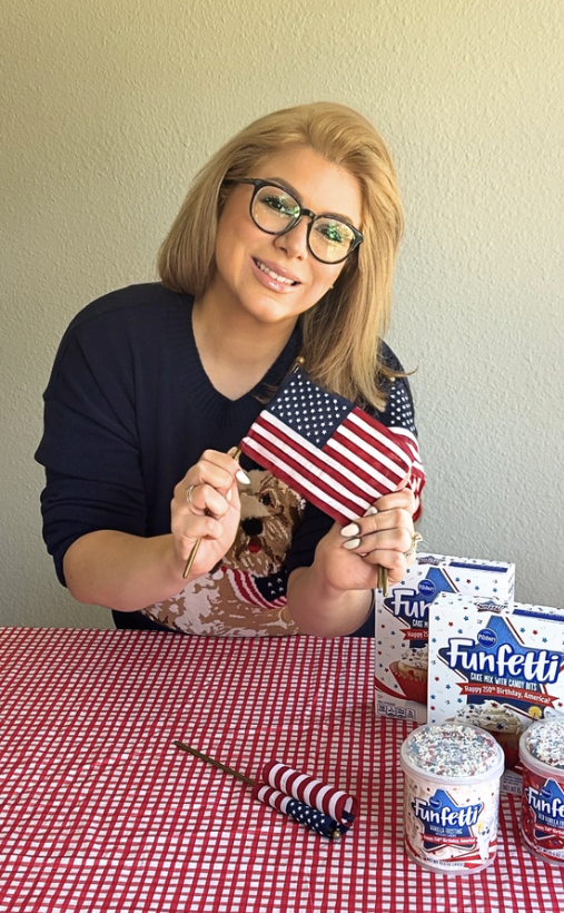 Woman smiling while holding two American flags at a red gingham table decorated with classic American snack cakes for a Hosting the World USA celebration.