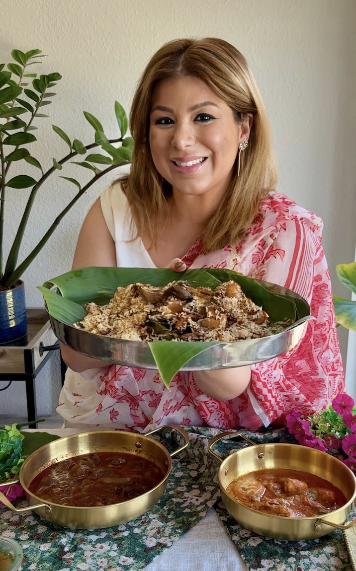 Smiling woman in a pink and white sari holding a large tray of authentic Bangladeshi kacchi biryani served on banana leaves, with traditional beef bhuna and chicken roast displayed on the table.