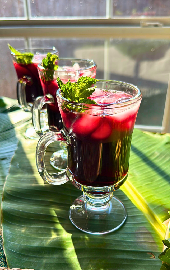 Glass mugs filled with traditional Senegalese bissap, a refreshing hibiscus drink served with ice and mint on a banana leaf table setting in natural sunlight.