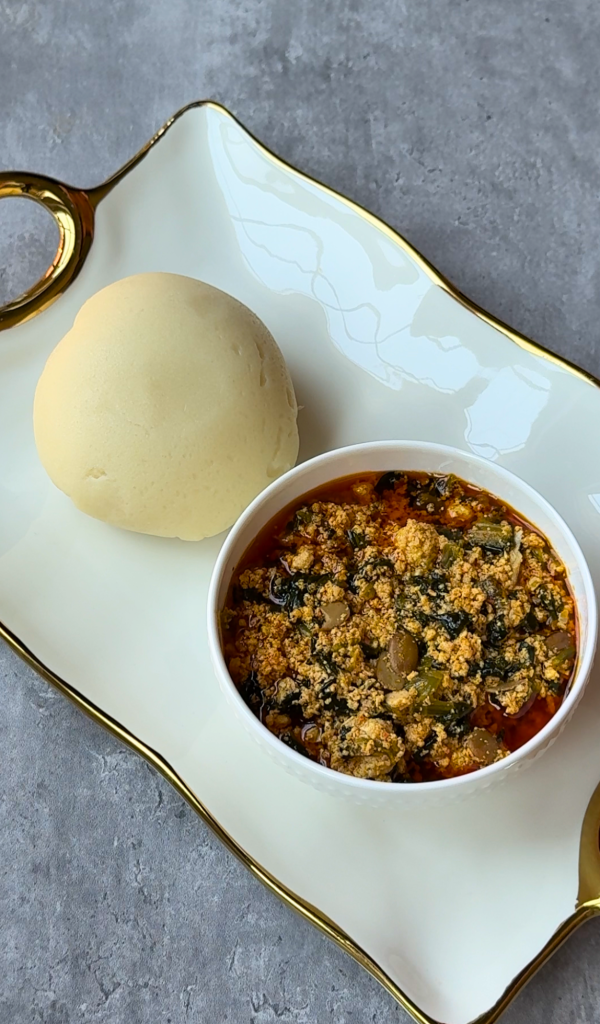 Traditional Nigerian egusi soup served in a white bowl with a ball of smooth fufu on a white serving platter.