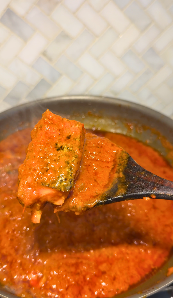 A piece of fish coated in rich Nigerian tomato stew is held on a wooden spoon above a pan of simmering fish stew.