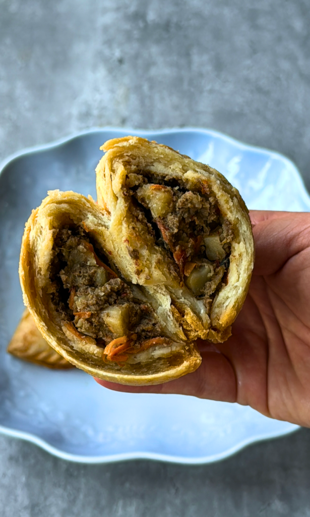 A Nigerian beef patty cut in half to reveal a savory filling of seasoned ground beef and diced potatoes inside a flaky golden pastry.