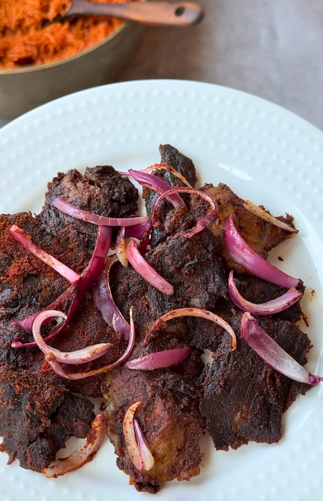 Homemade Nigerian beef suya served on a white plate with sliced red onions and traditional yaji spice, with jollof rice in the background.