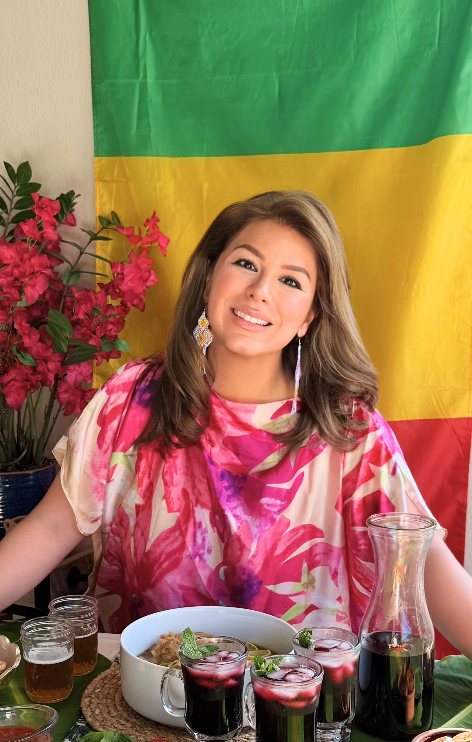 Woman wearing a pink Senegalese-inspired outfit seated behind a colorful Senegalese dinner table featuring thieboudienne, bissap, fresh salads, breads, and traditional West African dishes in front of a Senegal flag.